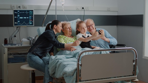 Niño tomando selfie en el teléfono inteligente con el paciente enfermo y la familia en la visita a la cama de la sala del hospital. Niño usando teléfono móvil para fotos y visitando a una anciana con un anciano y su madre en la clínica.