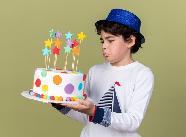 Niño triste con gorro de fiesta azul sosteniendo y mirando el pastel
