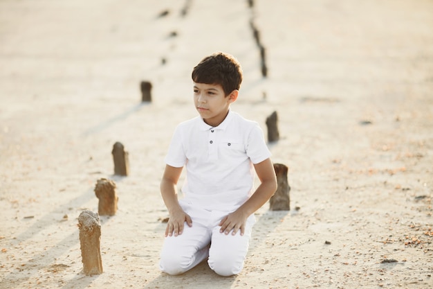 Foto gratuita niño vestido con ropa blanca está sentado de rodillas en la arena al atardecer