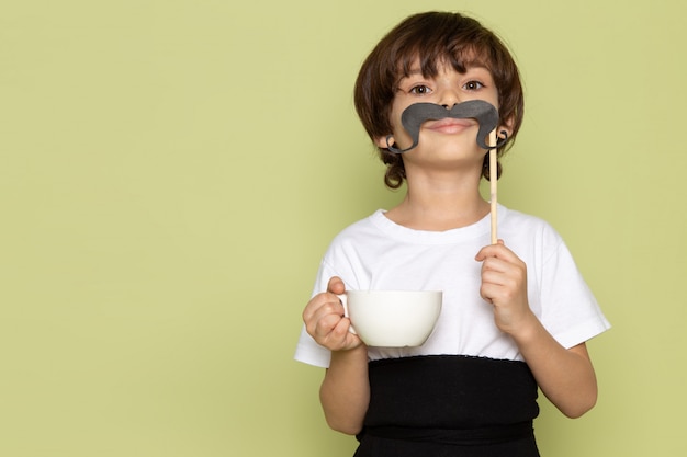 Un niño de vista frontal con bigote y taza de café en camiseta blanca en el piso de color piedra