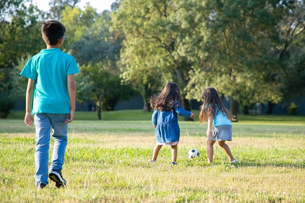 Niños activos jugando al fútbol en el césped en el parque de la ciudad. Vista trasera completa. Concepto de actividad infantil y al aire libre.