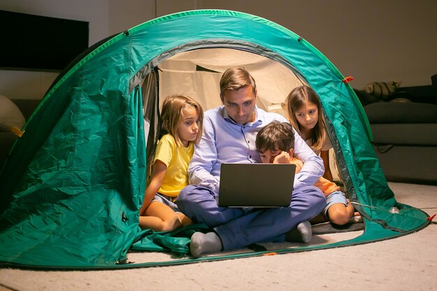 Niños adorables que se relajan con el padre en la tienda de campaña en casa y ven películas en la computadora portátil. Niños felices y papá amoroso sentado en la tienda con luz. Concepto de infancia, tiempo en familia y fin de semana.