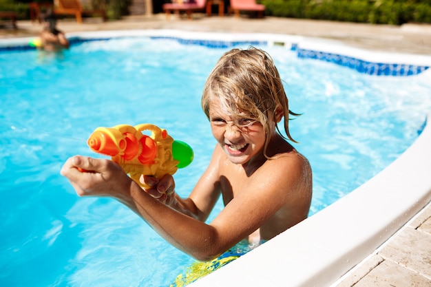 Niños alegres jugando pistolas de agua, regocijándose, saltando, nadando en la piscina.