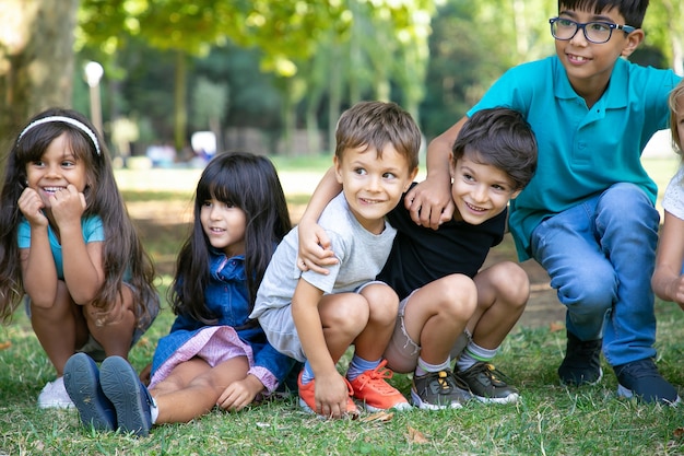 Niños alegres sentados y en cuclillas sobre el césped, abrazándose unos a otros, mirando a otro lado con emoción. Concepto de juego o entretenimiento para niños.