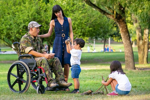 Niños arreglando madera para fogatas en el parque cerca de mamá y papá militar discapacitado en silla de ruedas. Niño mostrando registro al padre emocionado. Veterano discapacitado o concepto de familia al aire libre