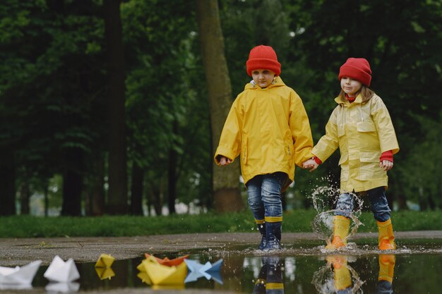 Niños divertidos en botas de lluvia jugando con barco de papel por un charco