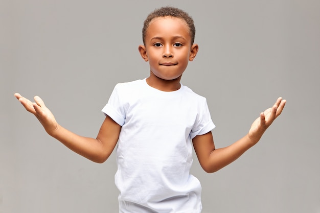 Niños, estilo de vida y lenguaje corporal. Disparo aislado de fresco y guapo niño afroamericano con mirada segura mordiendo el labio inferior y haciendo gestos con las palmas, mostrando que no tiene miedo