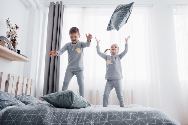 Niños felices jugando en el dormitorio blanco. Niño y niña, hermano y hermana juegan en la cama con pijama. Ropa de dormir y ropa de cama para bebés y niños pequeños. Familia en casa