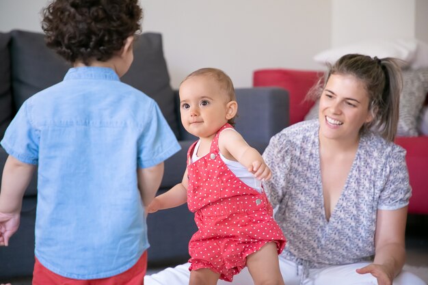 Niños felices jugando en la sala de estar con mamá rubia. Niña linda de pie y mirando al chico rizado. Madre cariñosa sonriente viendo a los niños. Concepto de familia en el interior, fin de semana y maternidad