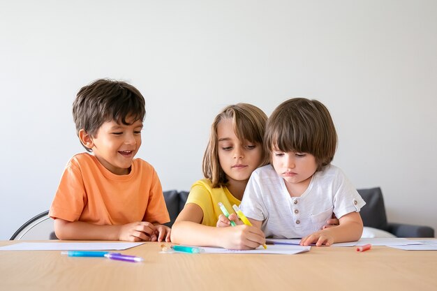 Niños felices pintando con rotuladores en la sala de estar. Niños encantadores y niña rubia sentados en la mesa, dibujando en papel con bolígrafos y jugando en casa. Concepto de infancia, creatividad y fin de semana.