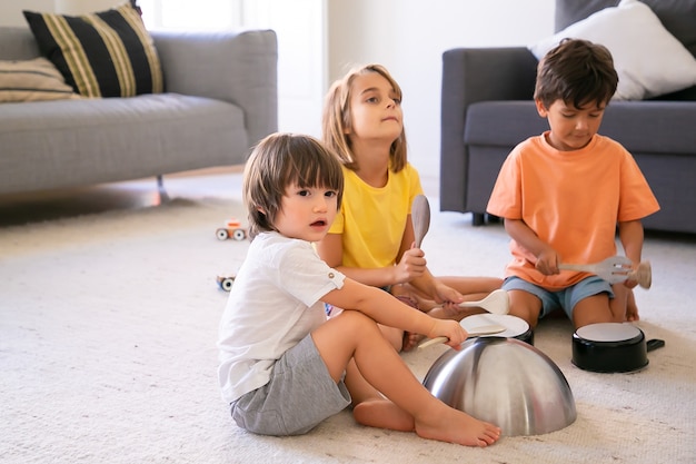 Niños felices sentados en la alfombra y jugando con utensilios. Lindos niños caucásicos y chica rubia divirtiéndose juntos en la sala de estar y golpeando sartenes. Concepto de actividad de la infancia y el hogar.