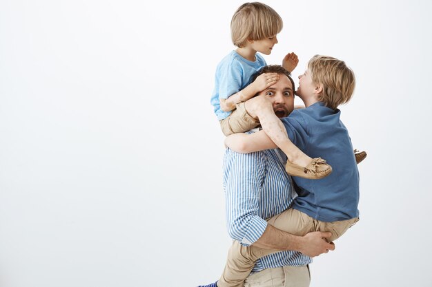 Niños jugando con papá genial. Retrato de juguetones hijos felices colgando del cuerpo del padre, divirtiéndose y jugando juntos