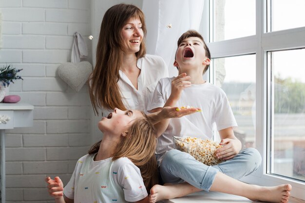 Niños juguetones comiendo palomitas de maíz con su madre en casa.