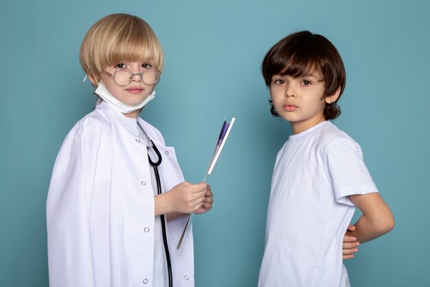 Niños lindos adorables mirando a la cámara, uno en traje de médico blanco y otro en camiseta blanca en la pared azul