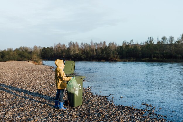 Los niños con las manos en los guantes recogiendo botellas vacías de plástico en una bolsa de basura cerca del lago