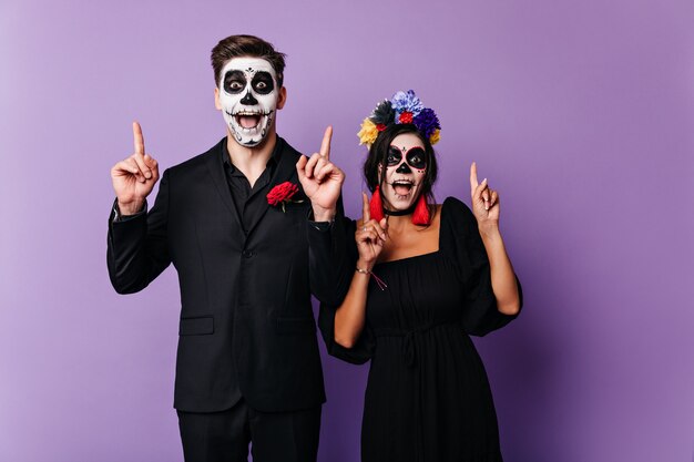 Los niños y niñas alegres de ojos oscuros posan emocionalmente, mostrando los dedos hacia arriba. Foto de pareja con arte de la cara en estilo mexicano en la pared púrpura.
