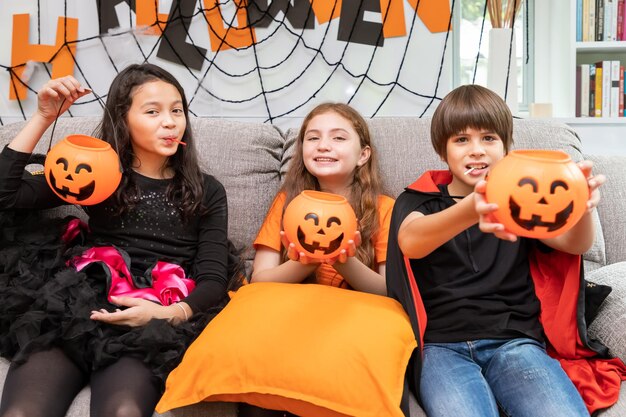 Niños y niñas felices y lindos disfrazados en un sofá en la sala de estar durante la fiesta de Halloween sosteniendo calabaza Jacko'lantern en la mano