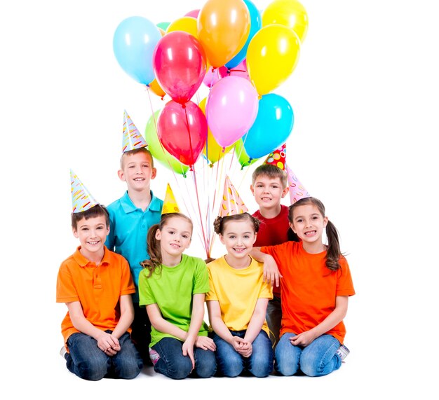 Niños y niñas felices con sombrero de fiesta con globos de colores sentados en el suelo - aislados en blanco