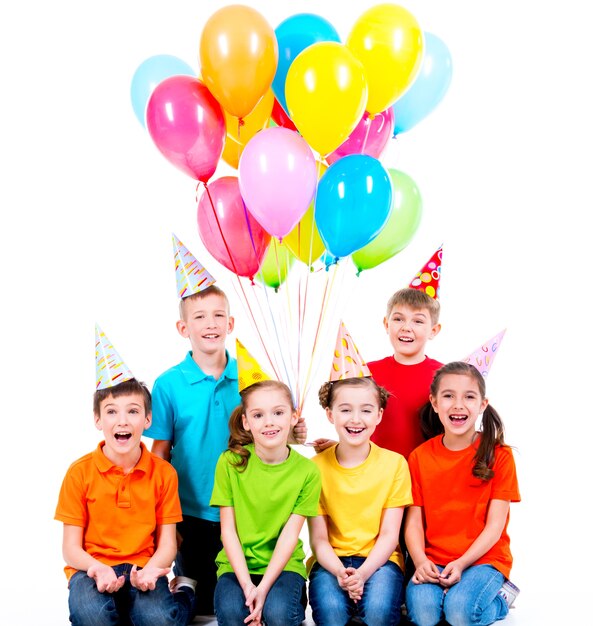 Niños y niñas felices con sombrero de fiesta con globos de colores sentados en el suelo - aislados en blanco
