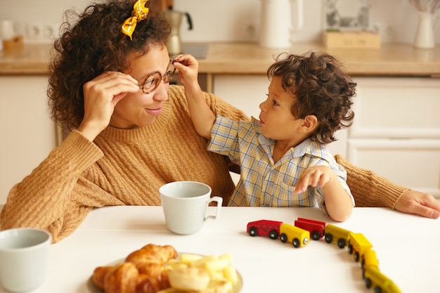 Niños, niños, infancia feliz, vínculos familiares y concepto de crianza. imagen de la atractiva joven hispana tomando un café en la mesa de ktichen y sonriendo mientras el hijo pequeño se quita las gafas