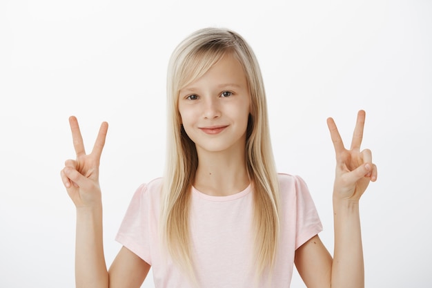 Niños de la paz. Chica joven alegre y emotiva con actitud positiva, sonriendo amistosamente y mostrando signos de victoria o v con ambas manos, haciendo fotos para la competencia de los niños sobre una pared gris