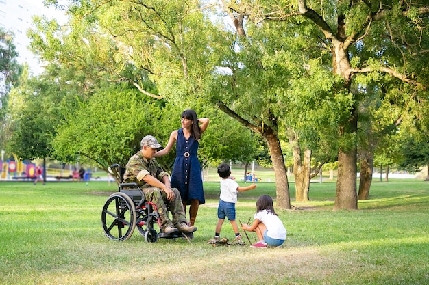 Niños pequeños arreglando leña para fogatas al aire libre cerca de mamá y papá militar discapacitado en silla de ruedas. Veterano discapacitado o concepto de familia al aire libre