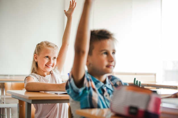 Niños que se sientan en los escritorios de la escuela que levantan las manos