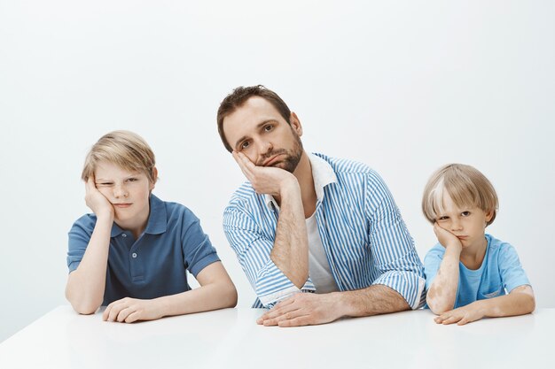 Los niños se sienten aburridos y molestos. Retrato de familia europea divertida cansada de hijos y papá sentados en la mesa, con la cabeza apoyada en las palmas y mirando indiferente