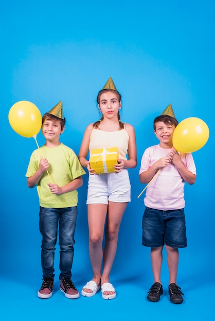 Niños en el sombrero de cumpleaños con regalo amarillo y globos amarillos contra el fondo azul