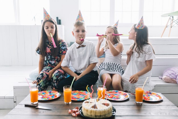 Niños con sopladores de fiesta cerca de pastel de cumpleaños y bebidas