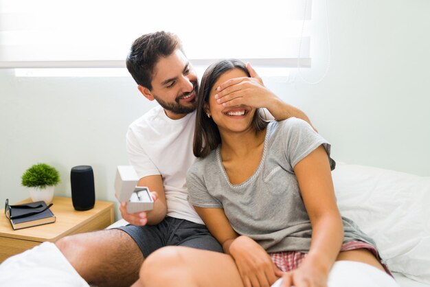 No abras los ojos. Hombre romántico enamorado cubriendo los ojos de su novia para sorprenderla con un anillo de compromiso después de despertarse juntos en la cama