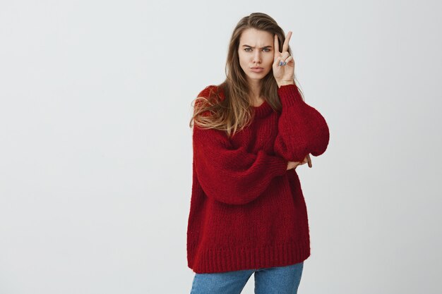 No es una muñeca para sonreír todo el tiempo. Foto de estudio de elegante mujer guapa en suéter suelto que muestra v signo de victoria o paz cerca de la cara mientras frunce el ceño y expresa disgusto u ofensa, de pie