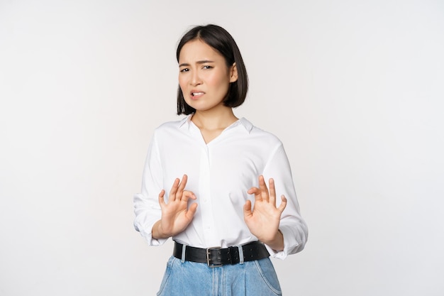 No, gracias Joven mujer asiática disgustada rechazando la propuesta dándose la mano y retrocediendo mirando con desagrado rechazando la oferta de pie sobre fondo blanco