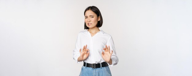 No, gracias Joven mujer asiática disgustada rechazando la propuesta dándose la mano y retrocediendo mirando con desagrado rechazando la oferta de pie sobre fondo blanco