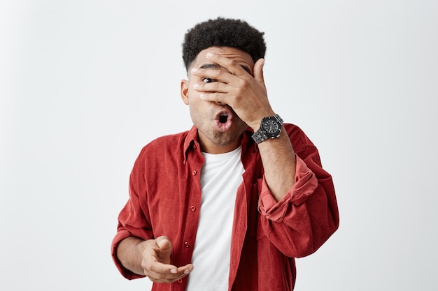 No hagas eso. Copia espacio Retrato de hombre joven de piel negra con peinado afro en camiseta blanca y camisa roja cara cercana con la mano, no puede mirar a su amigo haciendo estupideces.