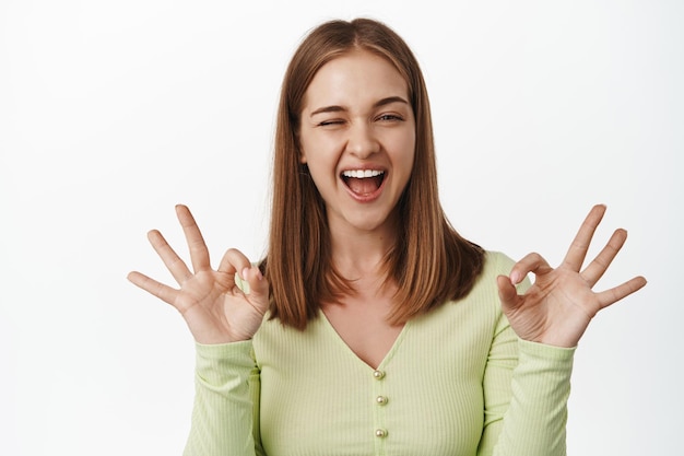 Foto gratuita no hay problema, está bien. la niña feliz guiña un ojo y sonríe, muestra el signo ok, cero ok, asegurando que todo esté bajo control, garantice la calidad del servicio, recomiende el lugar, de pie contra el fondo blanco.