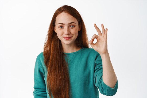 No hay problema, muy bien Una joven sonriente con el pelo largo y rojo se ve satisfecha, hacer bien, firmar cero, aprobar algo bueno y recomendar un fondo blanco.