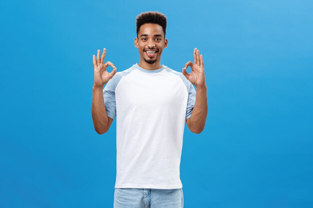 No hay problema. Retrato de alegre optimista novio de piel oscura con un corte de pelo fresco en una camiseta casual que muestra un gesto bien que le gusta la gran idea de un amigo sonriendo encantado posando sobre fondo azul