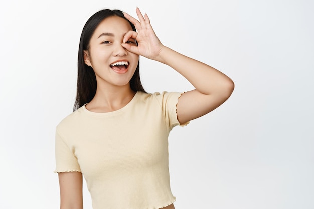 No hay problema Sonriente mujer asiática feliz mostrando cero gesto bien en el ojo riendo despreocupada de pie sobre fondo blanco.