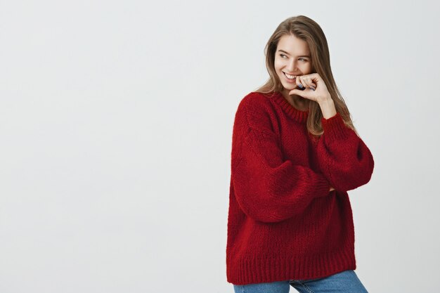 No hay tiempo para hablar en serio. Foto de estudio de emotiva chica juguetona europea en traje de moda con el dedo en el labio mientras mira a un lado en el espacio de la copia y sonríe sensual, coqueteando sobre la pared gris