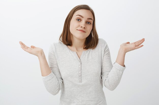 No me importa. Retrato de despreocupada mujer guapa complacida y tranquila en traje de moda encogiéndose de hombros inclinando la cabeza y sonriendo descuidado siendo desorientado y desinteresado sobre la pared blanca