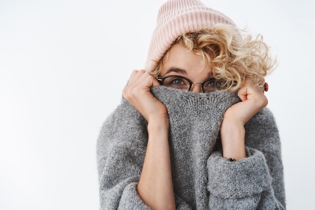 Foto gratuita no puedes conseguirme el invierno. linda chica rubia tonta y juguetona con gafas y gorro tirando del cuello del suéter en la cara escondiéndose y mirando con alegría, divirtiéndose jugando en la pared blanca.