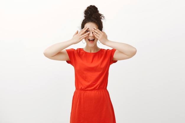 No puedo esperar a ver la sorpresa. Mujer emocionada positiva en elegante vestido rojo, sonriendo y cubriendo los ojos con las palmas, mirando a través de los dedos