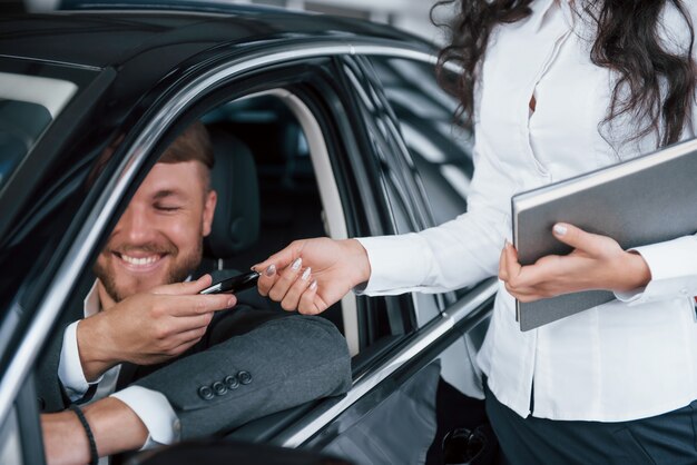 No puedo manejar las emociones. Dueño feliz de auto nuevo sentado adentro y toma las llaves de la gerente