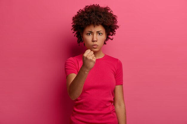 No te metas con mi ira. Mujer de piel oscura molesta aprieta el puño y parece irritada, insatisfecha con algo, amenaza con vengarse, usa camiseta casual, aislada en la pared rosa.