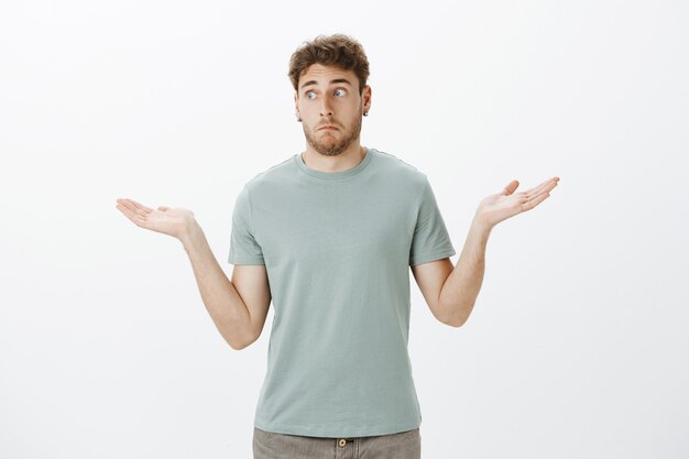 No tengo idea de cómo sucedió. Retrato de chico gracioso inconsciente incómodo en camiseta y aretes, haciendo una expresión despistada y encogiéndose de hombros con las palmas abiertas