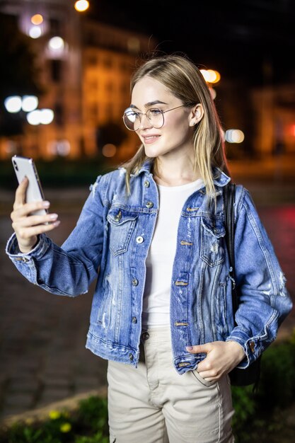 Noche al aire libre de la mujer joven con la cara del teléfono inteligente iluminada por la luz de la pantalla. Internet, red social, concepto de tecnología.