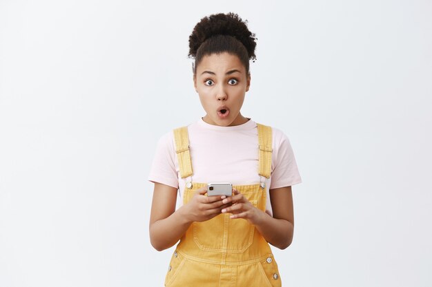 Noticias increíbles, compruébalo. Retrato de mujer afroamericana linda impresionado sorprendido en peto de moda amarillo, labios plegables mirando asombrado, sosteniendo el teléfono celular sobre la pared gris