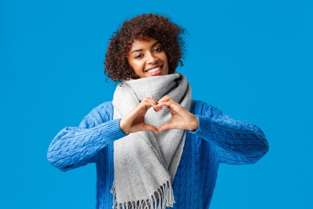 Novia afroamericana alegre encantadora, romántica con corte de pelo afro, inclinación de la cabeza mostrando el signo del corazón, confesando amor y afecto, con bufanda de invierno, suéter, de pie sobre la pared azul