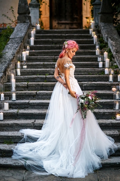 La novia con cabello rosado y tatuajes en su hombro tierno se encuentra sobre pisadas con velas brillantes